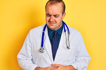 Young doctor man wearing coat and stethoscope standing over isolated yellow background with hand on stomach because indigestion, painful illness feeling unwell. Ache concept.