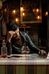 Drinking man. Man with beard holds glass brandy. Man holding a glass of whisky. Sipping whiskey. Degustation, tasting. Macho is drinking whiskey by his laptop.Thinking man. Tired.