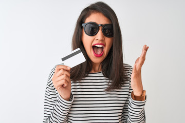 Naklejka premium Young chinese woman wearing sunglasses holding credit card over isolated white background very happy and excited, winner expression celebrating victory screaming with big smile and raised hands