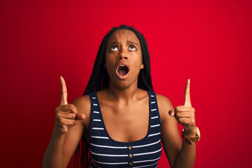 Young african american woman wearing striped t-shirt standing over isolated red background amazed and surprised looking up and pointing with fingers and raised arms.