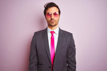 Young handsome businessman wearing suit and sunglasses over isolated pink background with serious expression on face. Simple and natural looking at the camera.