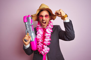 Young businessman wearing water gun hawaiian lei hat glasses over isolated pink background annoyed and frustrated shouting with anger, crazy and yelling with raised hand, anger concept