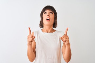 Young beautiful woman wearing casual t-shirt standing over isolated white background amazed and surprised looking up and pointing with fingers and raised arms.