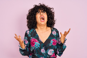 Young arab woman with curly hair wearing floral dress over isolated pink background crazy and mad shouting and yelling with aggressive expression and arms raised. Frustration concept.
