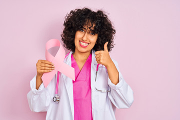 Young arab doctor woman with curly hair holding cancer ribbon over isolated pink background happy with big smile doing ok sign, thumb up with fingers, excellent sign