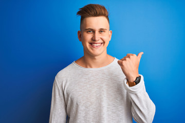 Young handsome man wearing white casual t-shirt standing over isolated blue background pointing and showing with thumb up to the side with happy face smiling