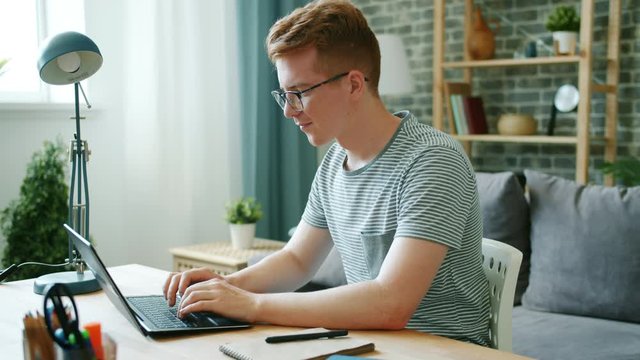 Teenage freelancer redhead young man is working with laptop at home typing smiling sitting at desk alone. Communication, occupation and people concept.