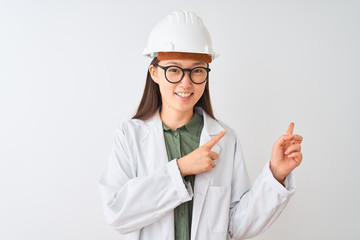 Young chinese engineer woman wearing coat helmet glasses over isolated white background smiling and looking at the camera pointing with two hands and fingers to the side.