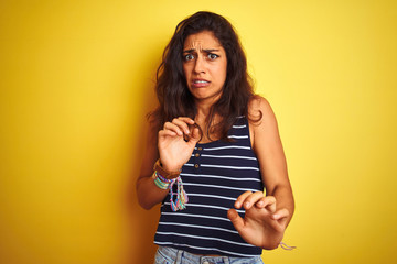 Young beautiful woman wearing striped t-shirt standing over isolated yellow background disgusted expression, displeased and fearful doing disgust face because aversion reaction. With hands raised