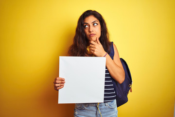 Naklejka na ściany i meble Young beautiful student woman holding banner standing over isolated yellow background serious face thinking about question, very confused idea