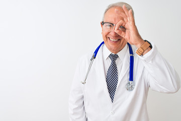 Senior grey-haired doctor man wearing stethoscope standing over isolated white background doing ok gesture with hand smiling, eye looking through fingers with happy face.