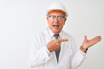 Senior grey-haired engineer man wearing coat and helmet over isolated white background amazed and smiling to the camera while presenting with hand and pointing with finger.