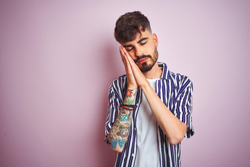 Fototapeta premium Young man with tattoo wearing striped shirt standing over isolated pink background sleeping tired dreaming and posing with hands together while smiling with closed eyes.