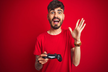 Young gamer man with tattoo playing video game standing over isolated red background very happy and excited, winner expression celebrating victory screaming with big smile and raised hands