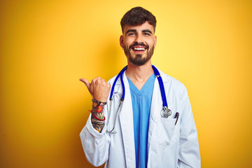 Young doctor man with tattoo wearing stethocope standing over isolated yellow background smiling...