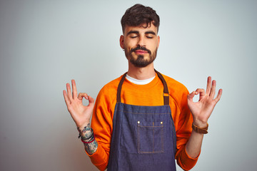 Young shopkeeper man with tattoo wearing apron standing over isolated white background relax and smiling with eyes closed doing meditation gesture with fingers. Yoga concept.