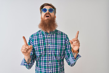 Young redhead irish man wearing casual shirt and sunglasses over isolated white background amazed and surprised looking up and pointing with fingers and raised arms.