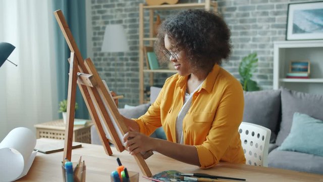 Beautiful African American girl is painting at home with pencil and paints sitting at desk alone. Creative young people, modern lifestyle and hobby concept.