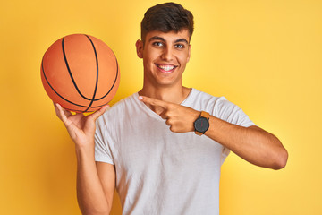 Young indian sportsman holding basketball ball standing over isolated yellow background very happy pointing with hand and finger