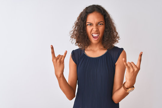 Young brazilian woman wearing blue dress standing over isolated white background shouting with crazy expression doing rock symbol with hands up. Music star. Heavy concept.