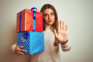 Young beautiful woman holding birthday gifts standing over isolated white background with open hand doing stop sign with serious and confident expression, defense gesture