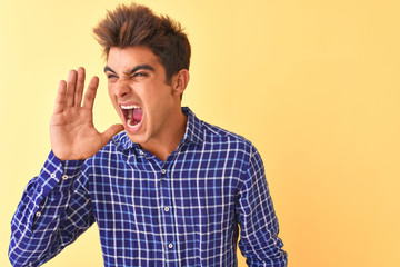 Young handsome man wearing casual shirt standing over isolated yellow background shouting and screaming loud to side with hand on mouth. Communication concept.