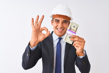 Young handsome architect man wearing helmet holding dollars over isolated white background doing ok sign with fingers, excellent symbol