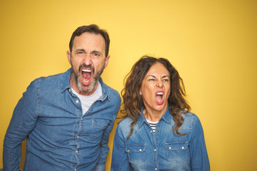 Beautiful middle age couple together standing over isolated yellow background angry and mad screaming frustrated and furious, shouting with anger. Rage and aggressive concept.