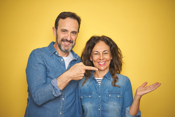 Obraz premium Beautiful middle age couple together standing over isolated yellow background amazed and smiling to the camera while presenting with hand and pointing with finger.