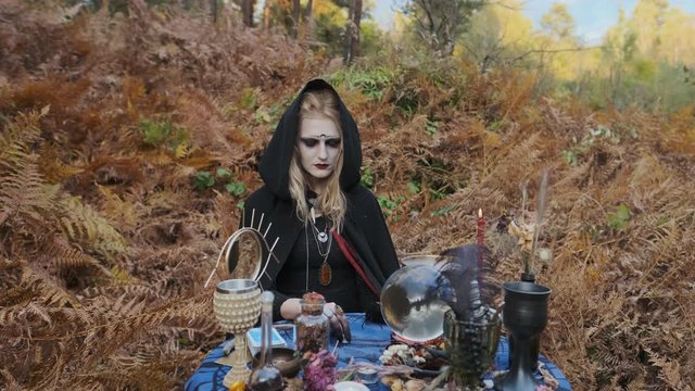 Young Witch Fortune-Tellers In The Forest. Witch At The Table Spreads The Magic Crystals. Halloween Image.