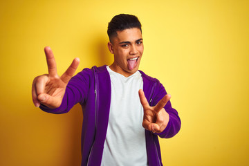 Young brazilian man wearing purple sweatshirt standing over isolated yellow background smiling with tongue out showing fingers of both hands doing victory sign. Number two.