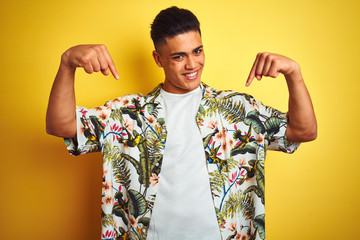 Young brazilian man on vacation wearing summer floral shirt over isolated yellow background looking...