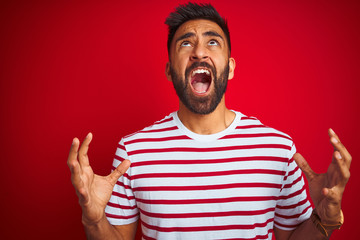 Young indian man wearing striped t-shirt standing over isolated red background crazy and mad shouting and yelling with aggressive expression and arms raised. Frustration concept.