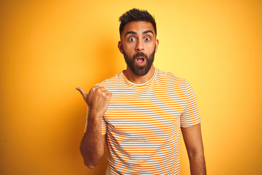 Young indian man wearing t-shirt standing over isolated yellow background Surprised pointing with hand finger to the side, open mouth amazed expression.