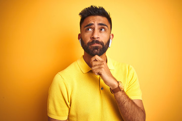 Young indian man listening to music using earphones standing over isolated yellow background with hand on chin thinking about question, pensive expression. Smiling with thoughtful face. Doubt concept.