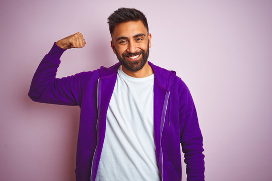 Young indian man wearing purple sweatshirt standing over isolated pink background Strong person showing arm muscle, confident and proud of power