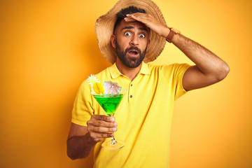 Indian man on vacation drinking cocktail wearing summer hat over isolated yellow background stressed with hand on head, shocked with shame and surprise face, angry and frustrated. Fear and upset