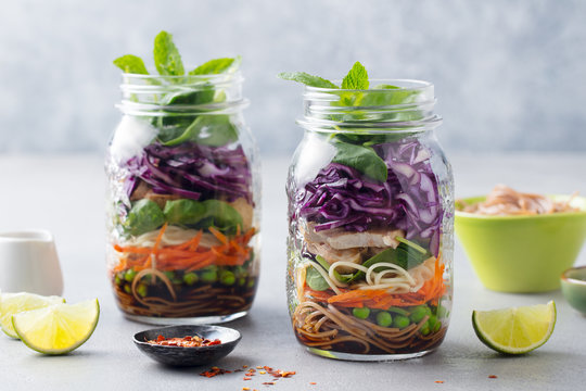 Healthy asian salad with noodles, vegetables, chicken and tofu in glass jars. Grey background.