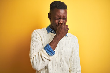 African american man wearing denim shirt and white sweater over isolated yellow background smelling something stinky and disgusting, intolerable smell, holding breath with fingers on nose