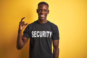 African american safeguard man wearing security uniform over isolated yellow background smiling and confident gesturing with hand doing small size sign with fingers looking and the camera