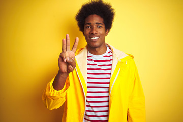 African american man with afro hair wearing rain coat standing over isolated yellow background showing and pointing up with fingers number three while smiling confident and happy.