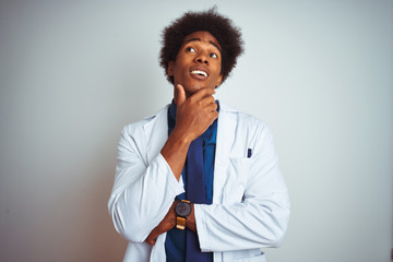 Young african american doctor man wearing coat standing over isolated white background with hand on chin thinking about question, pensive expression. Smiling with thoughtful face. Doubt concept.