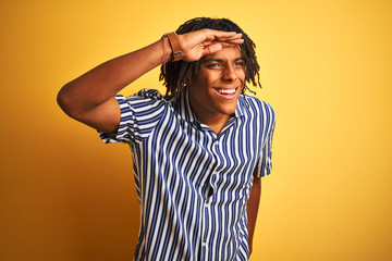 Afro man with dreadlocks wearing casual striped t-shirt over isolated yellow background very happy and smiling looking far away with hand over head. Searching concept.