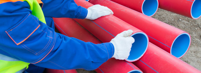 worker’s hands, in white gloves, against a background of red plastic pipes stacked on top of each...