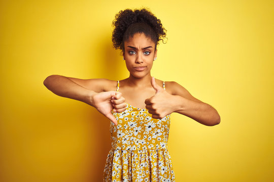 African american woman wearing casual floral dress standing over isolated yellow background Doing thumbs up and down, disagreement and agreement expression. Crazy conflict