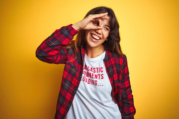 Beautiful woman wearing funny t-shirt with irony comments over isolated yellow background with happy face smiling doing ok sign with hand on eye looking through fingers
