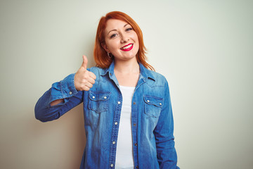 Young beautiful redhead woman wearing denim shirt standing over white isolated background doing happy thumbs up gesture with hand. Approving expression looking at the camera with showing success.