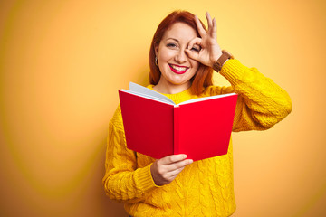 Young redhead teacher woman reading red book over yellow isolated background with happy face smiling doing ok sign with hand on eye looking through fingers