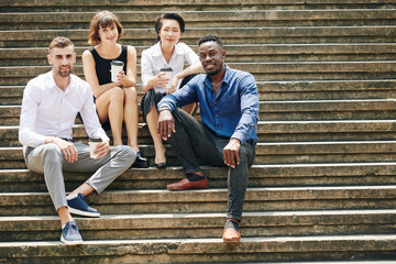 Cheerful business people resting on steps and enjoying coffee break on sunny day