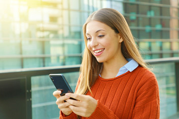 Young business woman wearing orange sweater texting on the smart phone out of the office in a sunny day.